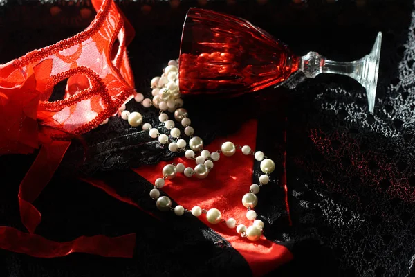 Uma máscara vermelha ao lado de uma taça de vinho tinto e um colar de pérolas, criando uma composição elegante.